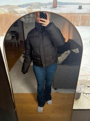 Svart pufferjacka med hög krage - Säljer en svart pufferjacka med hög krage och dragkedja framtill. Jackan har en boxig passform och quiltad design som ger en riktigt trendig look. Perfekt för kalla dagar när du vill hålla dig varm och snygg. Passar till det mesta i garderoben. Använd 1 gång 