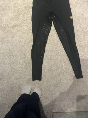 Svarta ridtights från Maya Delorez - Säljer ett par svarta ridtights från Maya Delorez i storlek XXS. Tightsen har silikonmönster på insidan av benen för extra grepp och en liten gul logga på låret. Tillverkade i stretchigt syntetmaterial som passar perfekt för ridning.