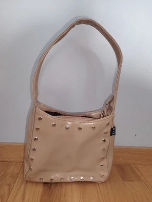 Beige handväska med nitar - Trendig beige handväska i fyrkantig modell med silverfärgade nitar på framsidan. Väskan har bred axelrem och dragkedja upptill. Tillverkad i ett slätt, konstläderliknande material. Perfekt för dig som gillar en edgy men stilren look. Är i fint använt använt skick, det enda att anmärka är att den skrapats lite på sidan av axelremmen (se bild). 