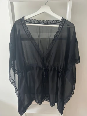 Svart transparent loungewear med spets - Svart transparent loungewear från Lindex med v-ringning och dekorativa spetsdetaljer längs kanterna. Plagget har korta ärmar och knytband i midjan för en avslappnad passform. Kan även användas som beachware nu till sommaren. 