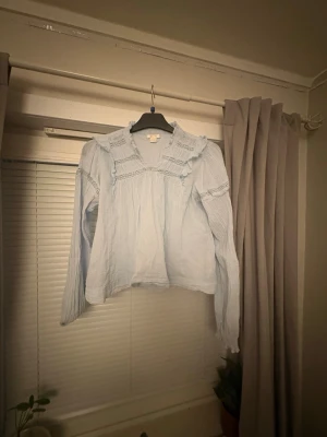 Ljusblå volangblus från Gina Tricot - Supersöt ljusblå blus från Gina Tricot med volangdetaljer och spets längs axlar och bröst. Blusen har v-ringning och långa ärmar i ett tunt, luftigt tyg som ger en romantisk vibe. Perfekt att styla med jeans eller kjol för en trendig look.