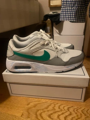 Nike Air Max sneakers - Nike Air Max sneakers i vitt och grått med gröna swoosh-detaljer. Skorna har en klassisk låg siluett, synlig Air-enhet i sulan och är tillverkade i en mix av mesh och syntetmaterial. Perfekta för dig som gillar retrostil och bekväma sneakers.