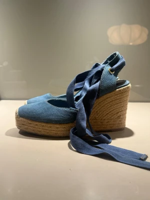 Blå kilklackade espadrilles med snörning - Säljer ett par blå espadrilles med kilklack och långa band att knyta runt ankeln. Ovandelen är i denimtyg och sulan är i flätad jute. Nästan aldrig använda!