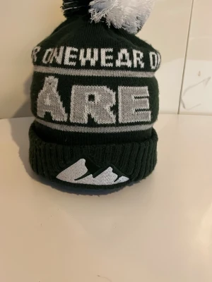 Mörkgrön Åre mössa med tofs - Stickad mörkgrön mössa från Are One Wear med stor vit text 'ÅRE' och vit tofs på toppen. Upprullad kant med broderad bergslogga framtill. Perfekt för kalla dagar och vintersäsongen.