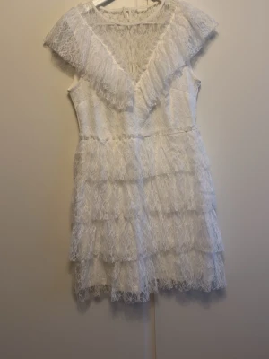 Vit spetsklänning med volanger - Söt vit kortklänning med flera lager volanger och spetsdetaljer. Klänningen har v-ringning och är ärmlös, vilket ger en romantisk och luftig känsla. Perfekt för sommarens alla tillfällen.