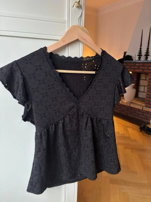 Svart blus med spets och volang - Söt svart blus med v-ringning, spetskant och volangärmar. Blusen har ett broderat hålmönster och är tillverkad i ett lätt bomullstyg. Perfekt för dig som gillar romantiska detaljer och en luftig känsla.