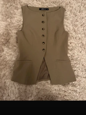 Beige väst från Gina Tricot - Stilren beige väst från Gina Tricot med knappar framtill och två dekorativa fickor. Västen har en figurnära passform och har en snygg och modern look. Perfekt att styla över en skjorta eller t-shirt.