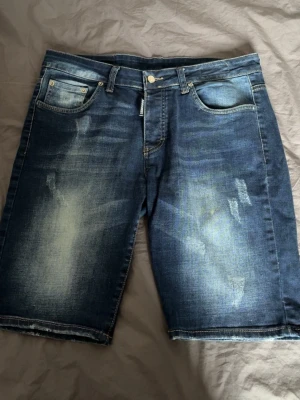 Blå jeansshorts från Dsquared2 - Säljer ett par snygga blå jeansshorts från Dsquared2 med slitna detaljer och klassisk femficksmodell. Shortsen har en cool tvättad look, knappgylf och kontrastsömmar. Perfekta för en avslappnad streetwear-stil.