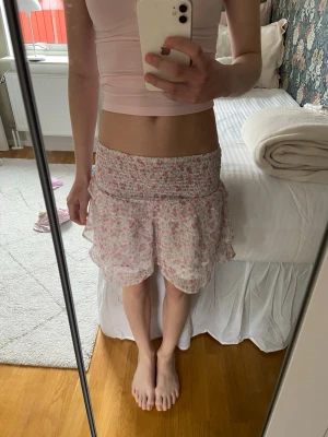 Blommig volangkjol i rosa och vit - Supersöt kort kjol med blommigt mönster i rosa och vitt. Kjolen har bred smock i midjan och dubbla volanglager för en luftig och lekfull känsla. Perfekt för varma dagar och enkel att matcha med olika toppar.