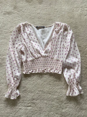 Prickig topp med volangärm - Söt topp i ljusrosa med lila prickar. Toppen har v-ringning, långa ärmar med volang vid ärmslut och smockad midja för en snygg croppad passform. Perfekt att styla med jeans eller kjol.