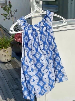 Blåvit mönstrad linnetopp - Säljer en luftig linnetopp i vitt med blått grafiskt mönster. Toppen har breda axelband med rynkdetalj och är ärmlös. Perfekt för varma dagar och ger en fräsch sommarkänsla.