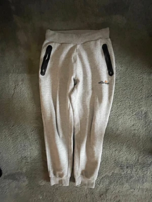 Grå mjukisbyxor från Ellesse - Snygga grå mjukisbyxor från Ellesse med svarta dragkedjor på fickorna och broderad logga på ena benet. Byxorna har resår i midjan och vid bensluten för en skön passform. Perfekta för chill eller träning.