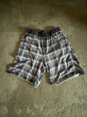 Rutiga pyjamasshorts i bomull - Säljer ett par rutiga shorts i svart, grått och vitt. De har elastisk midja och är tillverkade i mjuk bomull, perfekta för varma dagar. Passformen är loose och de har en avslappnad stil.
