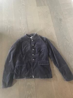 Mörkblå manchesterjacka med knappar - Stilren mörkblå jacka i manchester med markerad midja och dekorativa metallknappar framtill. Jackan har lång ärm, två fickor och en rak, något figurnära passform. Perfekt för dig som vill ha en klassisk men trendig look.