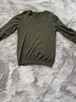 Olivgrön matinique tröja med rund hals - En enkel och stilren olivgrön tröja med rund halsringning och långa ärmar. Tröjan har ribbade muddar vid ärmslut och nederkant, perfekt för en clean och avslappnad look. Passar till det mesta och är lätt att matcha.