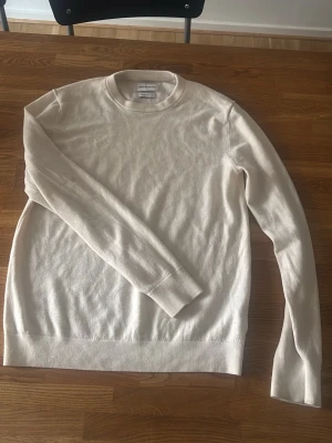 Beige merinoulltröja These Glory Days - Stilren beige stickad tröja från These Glory Days i mjuk superior merinoull. Tröjan har rund halsringning, långa ärmar och ribbade muddar vid ärmslut och nederkant. Perfekt för lager-på-lager och enkel att matcha.