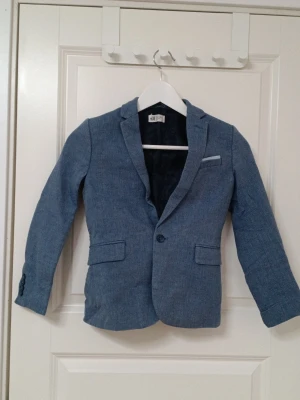 Blå kavaj från H&M barn storlek 134 - Snygg blå kavaj från H&M för barn. Kavajen har, en bröstficka och knappstängning. Fodrad insida och dekorativa knappar vid ärmslut. Perfekt för en dressad look. Storlek 134