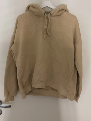 Beige hoodie från 157 med huva - Säljer en beige hoodie från 157 i storlek M. Hoodien har en stor huva med dragsnören och ribbade muddar vid ärmslut och nederkant. Materialet är mjukt och fleeceliknande på insidan, perfekt för att hålla sig varm. Enkel och stilren design.
