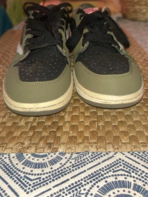 Nike Air Jordan 1 Low Travis Scott - Snygga Nike Air Jordan 1 Low Travis Scott sneakers i olivgrönt och svart med vita detaljer. Skorna har svarta skosnören, perforerad tåbox och röda broderade loggor på hälen. Tillverkade i läder och textil för en clean och modern look.