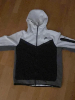 Nike svartvit vindjacka med huva - Nike vindjacka i svart och vitt med dragkedja framtill och huva. Jackan har en sportig design med kontrastpaneler och Nike-logga på bröstet. Tillverkad i lätt syntetmaterial som passar perfekt för blåsiga dagar.