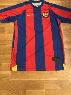 Barcelona fotbollströja Ronaldinho - Klassisk FC Barcelona matchtröja från Nike med röda och blå vertikala ränder, klubbmärke på bröstet och Ronaldinho 10 tryckt i gult på ryggen. Tillverkad i lätt och ventilerande material, perfekt för fotboll eller samling.