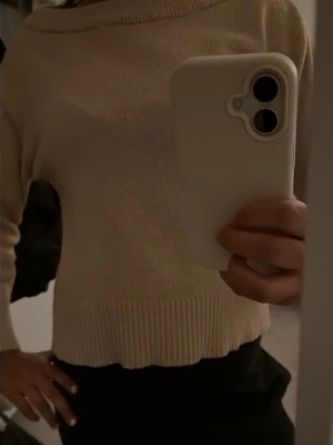 Beige stickad tröja med bred hals - Mysig beige stickad tröja med bred halsringning och ribbade muddar. Tröjan har en avslappnad passform och är perfekt att matcha med både jeans och kjol. Materialet ser ut att vara mjukt och skönt, troligen bomull eller en bomullsblandning.