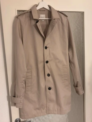 Beige trenchcoat från Premium by Jack & Jones - Stilren beige trenchcoat med klassisk krage, axelklaffar och svarta knappar. Jackan har raka fickor och justerbara ärmslut. Tillverkad i ett slätt bomullstyg som ger en clean look. Perfekt för dig som vill ha en tidlös och enkel trench.