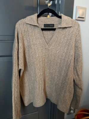 Beige kabelstickad tröja från ZARA - Snygg beige kabelstickad tröja från ZARA med krage och v-ringning. Tröjan har långa ärmar och en avslappnad passform, perfekt för lager på lager. Mjuk och skön kvalitet som passar till många olika stilar.