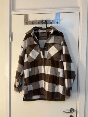 Brun och vit rutig overshirt - Overshirt i brun och vit rutigt mönster med stora bröstfickor och krage. Jackan har knappar framtill och raka ärmar. Perfekt för lager på lager och ger en avslappnad streetwear-look. Materialet ser ut att vara mjukt och lite tjockare, troligen bomull eller ullmix.