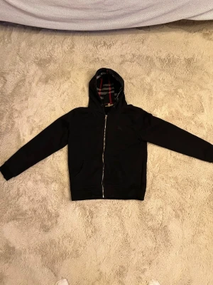 Burberry hoodie svart - Snygg svart hoodie från Burberry med dragkedja framtill och två fickor. Insidan av huvan har det klassiska rutmönstret och bröstet pryds av en diskret broderad logga. Perfekt för dig som gillar stilrena plagg med en twist.