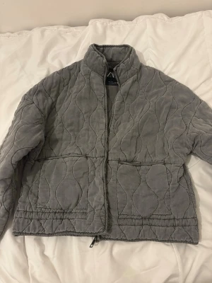 Grå quiltad pufferjacka från Zara - Säljer en grå quiltad pufferjacka från Zara som är slutsåld. Jackan har två stora fickor och ett vågigt mönster i tyget som ger en modern look. Perfekt för kalla dagar och passar bra nu till våren. 