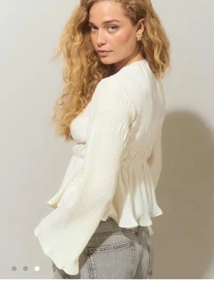Vit plisserad blus med v-ringning - Superfin vit blus med plisserat tyg och v-ringning. Modellen har markerad midja och volang nertill, samt långa ärmar som ger en romantisk vibe. Perfekt att styla med jeans eller kjol för en trendig look.