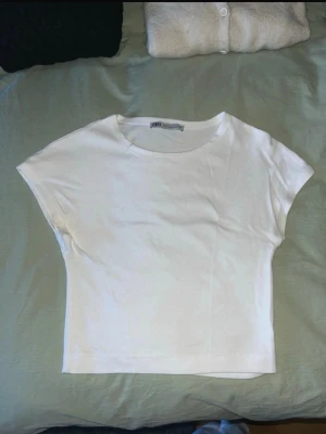Vit croppad topp från Zara - En enkel och stilren vit croppad topp från Zara. Toppen har korta ärmar och rund halsringning. Perfekt att matcha med höga jeans eller kjol för en clean look. Materialet känns mjukt och stretchigt.