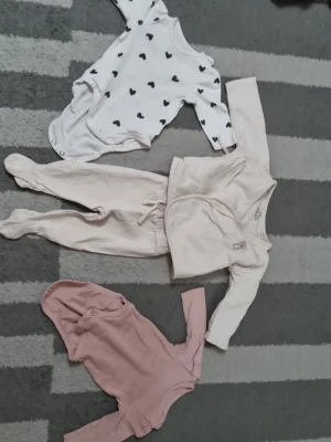 Sött babyklädpaket i neutrala färger - Paket med tre babyplagg: en vit långärmad body med svarta hjärtan, en ljusbeige omlottkofta med matchande byxa med fot samt en rosa långärmad body. Perfekt för mysiga dagar och enkla att matcha.