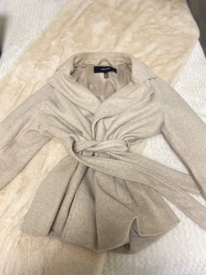 Beige kappa från Vero Moda med skärp - Stilren beige kappa från Vero Moda med bred krage och knytskärp i midjan. Kappan har en mjuk, ull-liknande yta och är helfodrad för extra komfort. Perfekt för dig som vill ha en klassisk och trendig look under kyliga dagar.Köpt för 750 säljs för 520 kr