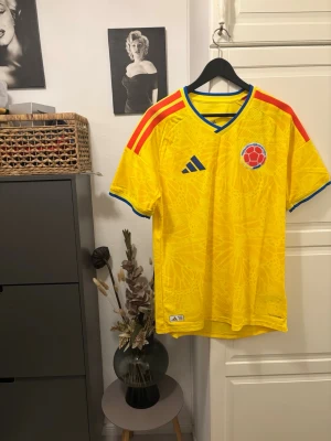 Gul Colombia fotbollströja (authentic) - Snygg gul Colombia fotbollströja från Adidas med röda och blå detaljer. Tröjan har korta ärmar, rund halsringning med blå kant och mönstrad struktur i tyget. Colombia-logga på bröstet och Adidas-logga i blått. Perfekt för match eller träning.