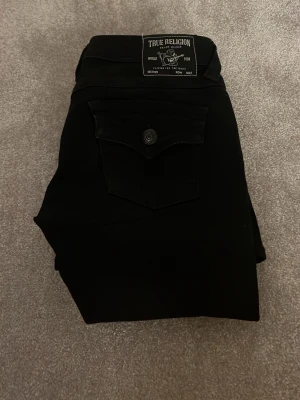 Svarta jeans från True Religion - Snygga svarta jeans från True Religion med klassisk femficksmodell och knappdetalj på bakfickan. Jeansen har en utsvängd passform och är tillverkade i ett mjukt bomullsmaterial med stretch för extra komfort. Perfekta för dig som gillar stilrena och tidlösa plagg.