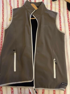 Grå softshellväst från Mountain Horse - Snygg grå softshellväst från Mountain Horse med vita dragkedjor och hög krage. Västen har två framfickor med dragkedja och en ficka bak. Perfekt för lager-på-lager och outdoor vibes.