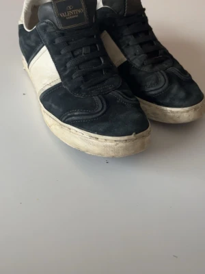 Svarta och vita sneakers från Valentino - Snygga sneakers från Valentino Garavani i svart mocka och vitt läder. Skorna har svarta snören, vit sula och coola detaljer på sulan med kamouflagemönster undertill. Perfekta för dig som vill ha en exklusiv och trendig look.