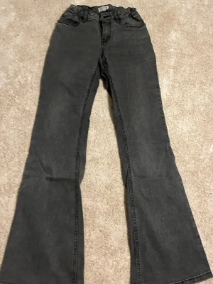 Grå utsvängda jeans från Lindex - Säljer ett par grå jeans från Lindex i modell med utsvängda ben. Jeansen har klassisk femficksdesign, hällor för bälte och dragkedjegylf. Perfekta för dig som gillar en retroinspirerad look och vill ha något bekvämt till vardags. Dom har en liten vit fläck på benet som man knappt ser.