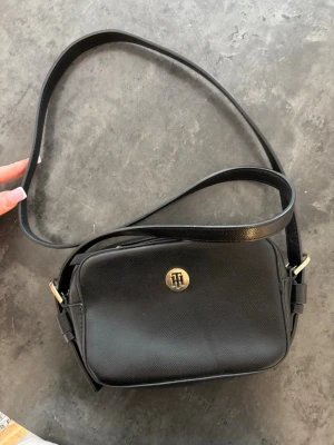 Svart axelväska från Tommy Hilfiger - Snygg svart axelväska i läderimitation från Tommy Hilfiger med guldfärgade detaljer och justerbar axelrem. Väskan har ett stilrent TH-emblem framtill och dragkedja upptill. Perfekt storlek för det viktigaste och passar till många outfits.