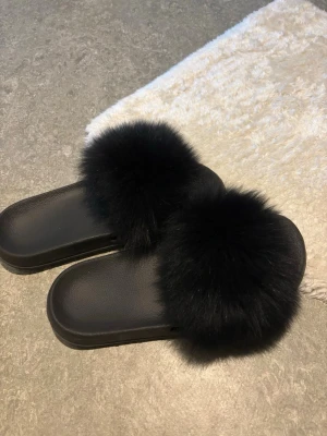 Svarta fluffiga tofflor med päls - Säljer ett par svarta tofflor med breda remmar täckta av mjuk, fluffig fuskpäls. Sulan är i svart syntetmaterial och har en enkel slip-in design. Perfekta för att chilla hemma eller ge din outfit en trendig touch.