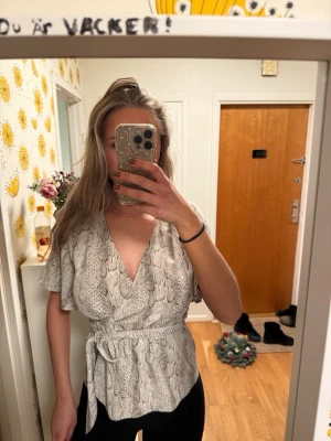 Grå omlottblus med ormmönster - Snygg omlottblus i grått med ormmönster och knytskärp i midjan. Blusen har korta, vida ärmar och djup v-ringning. 