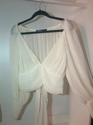 Plisserad vit omlottblus - Trendig vit omlottblus med plisserat tyg och djup v-ringning. Blusen har långa, luftiga ärmar och markerad midja som ger en snygg siluett. Perfekt för dig som gillar en stilren och modern look.