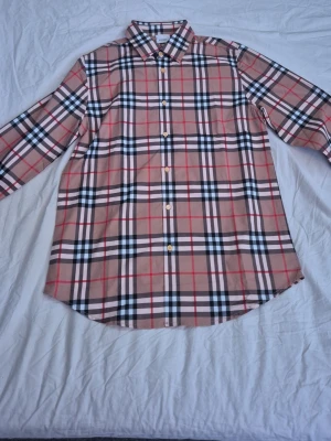 Rutig skjorta från Burberry - Klassisk rutig skjorta från Burberry i beige, svart, vit och röd. Skjortan har lång ärm, knappar framtill och en stilren krage. Tillverkad i bomull och har det ikoniska Burberry-mönstret. Perfekt för en trendig och tidlös look.