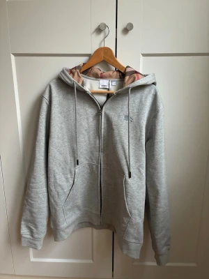 Grå zip hoodie från Burberry - Snygg grå zip hoodie från Burberry med klassiskt rutigt foder i huvan och diskret logga på bröstet. Perfekt för en chill och stilren look.