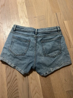 Blå jeansshorts med fransig kant - Säljer ett par klassiska blå jeansshorts från Lindex med fransig kant nertill och femficksmodell. Shortsen har bälteshällor och knappgylf. Perfekta för varma dagar och enkel att matcha med allt.