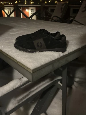 Svarta Fred Perry sneakers - Snygga svarta sneakers från Fred Perry med klassisk logga på sidan. Skorna har lågt skaft, svart ovandel i mocka och textil samt svarta snören. Perfekta för en stilren och avslappnad look.