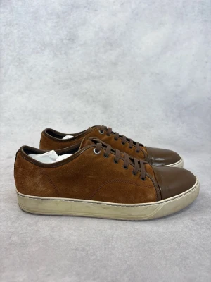 Bruna Lanvin sneakers i mocka och läder - Snygga bruna sneakers med ovandel i mocka och tåparti i blankt läder. Skorna har bruna skosnören och en vit sula med diskret struktur. Perfekta för dig som gillar en stilren och klassisk look med en twist.