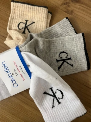 Calvin Klein 3-pack strumpor - Säljer ett 3-pack ribbade strumpor från Calvin Klein i vitt, grått och beige. Strumporna har svart CK-logga på sidan och svart kant upptill. Materialet är en blandning av bambu och bomull för extra komfort.